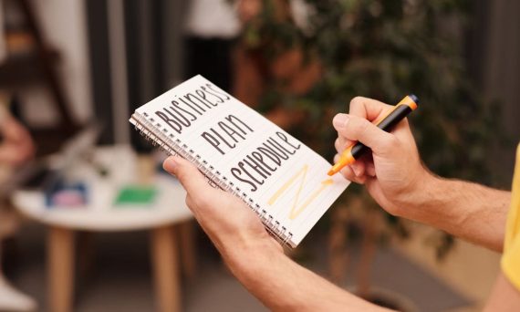 Close-up of a hand writing in a notebook with 'Business Plan Schedule' indoors.